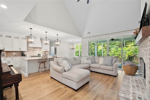 living room featuring high vaulted ceiling, a fireplace, light wood finished floors, and ceiling fan with notable chandelier