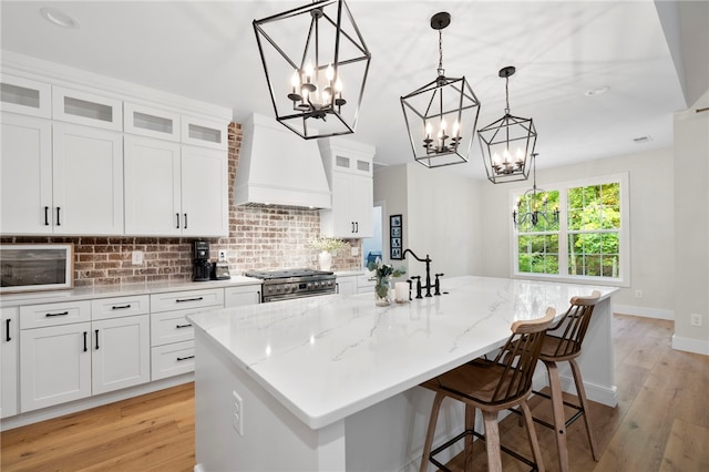 kitchen featuring custom exhaust hood, glass insert cabinets, white cabinetry, an island with sink, and high end range