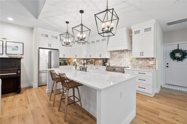 kitchen featuring a kitchen island with sink, stainless steel appliances, white cabinetry, glass insert cabinets, and custom range hood