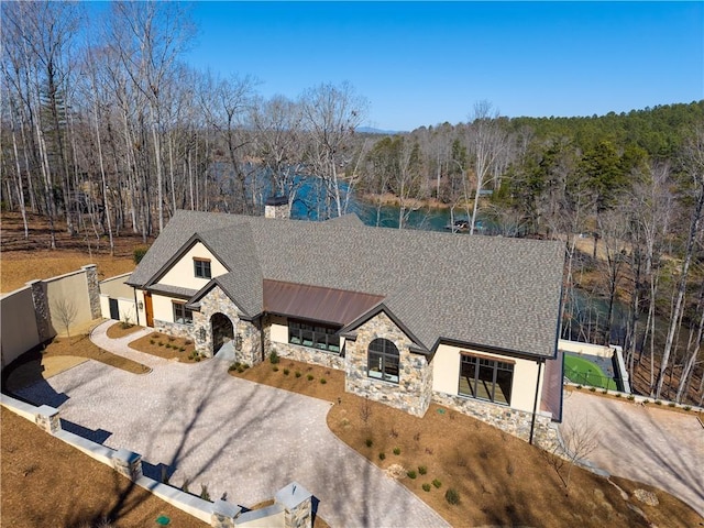 view of front facade featuring driveway, a shingled roof, a forest view, stone siding, and fence