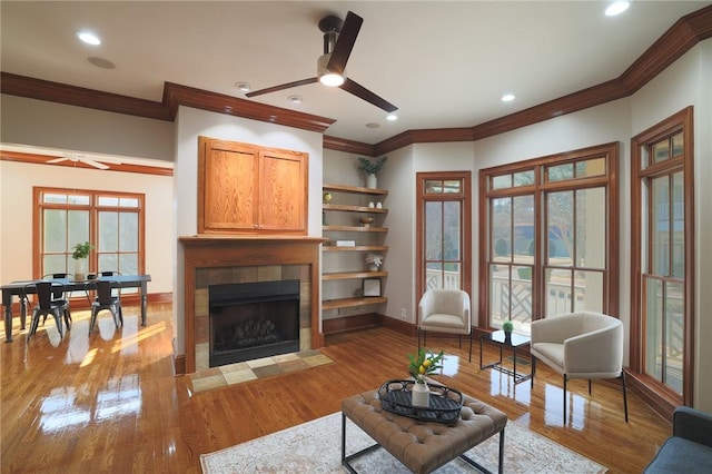 living room featuring a tiled fireplace, a ceiling fan, ornamental molding, wood finished floors, and baseboards