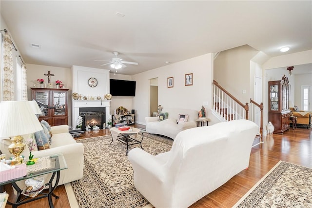 living room with light wood finished floors, visible vents, a ceiling fan, a warm lit fireplace, and stairs