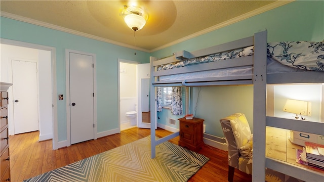 bedroom with wood finished floors, visible vents, baseboards, ornamental molding, and ensuite bath