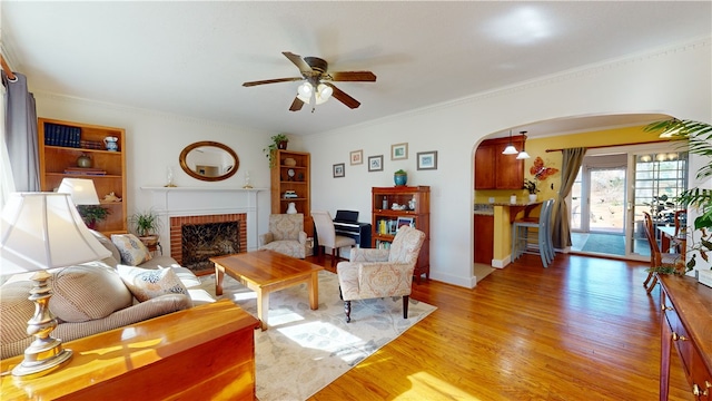 living area featuring arched walkways, a ceiling fan, light wood-style flooring, crown molding, and a brick fireplace