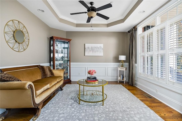 living area with a wainscoted wall, ceiling fan, ornamental molding, wood finished floors, and a tray ceiling