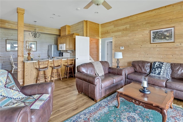 living room featuring ceiling fan with notable chandelier, light wood-type flooring, decorative columns, and wooden walls