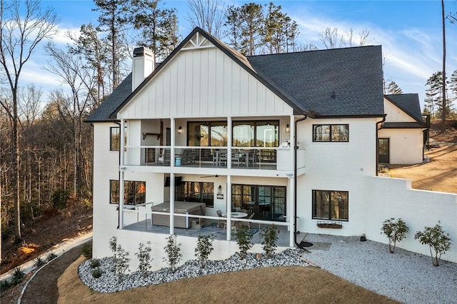 rear view of house with a patio, a balcony, roof with shingles, board and batten siding, and a chimney