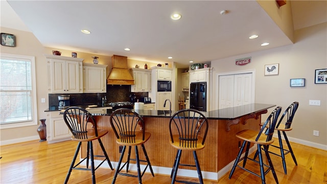 kitchen featuring black appliances, light wood finished floors, custom exhaust hood, and decorative backsplash