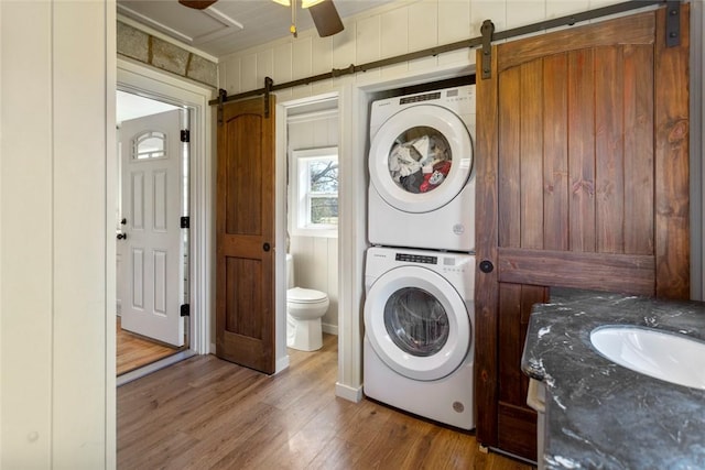 laundry area featuring laundry area, a barn door, a ceiling fan, stacked washer and clothes dryer, and wood finished floors