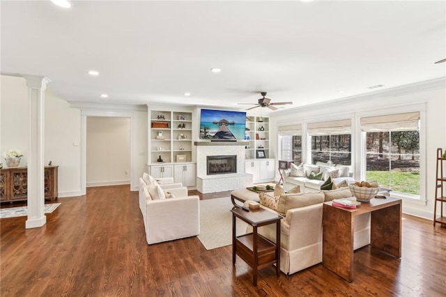 living area featuring dark wood-style floors, a fireplace with raised hearth, ceiling fan, and ornate columns