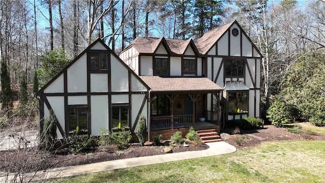 english style home with covered porch, a shingled roof, a front lawn, and stucco siding