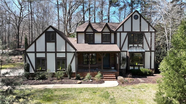 tudor-style house with a porch, brick siding, roof with shingles, stucco siding, and a front yard