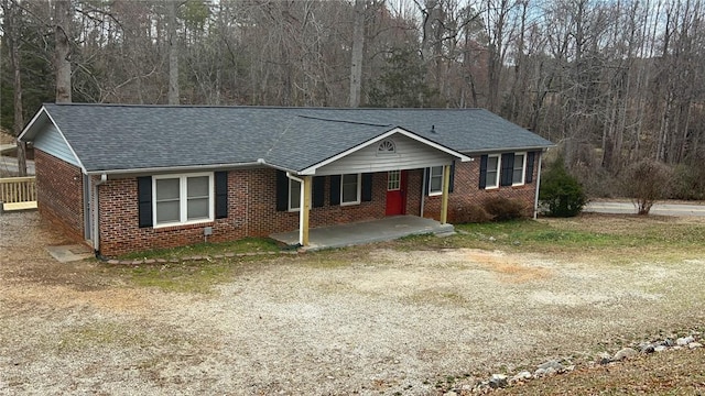 ranch-style home with driveway, a patio, a forest view, roof with shingles, and brick siding