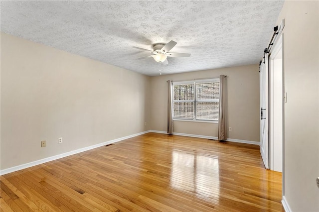 empty room with a textured ceiling, a barn door, a ceiling fan, baseboards, and light wood-type flooring