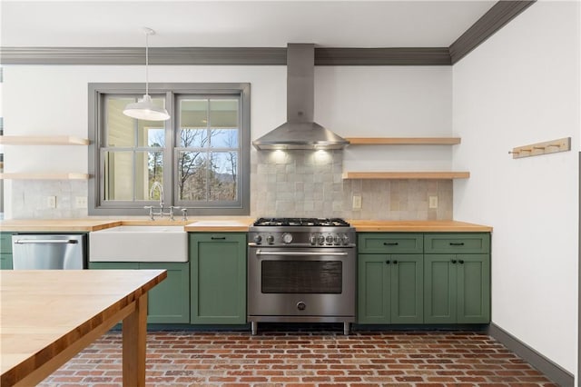 kitchen with wall chimney range hood, green cabinetry, butcher block counters, appliances with stainless steel finishes, and a sink