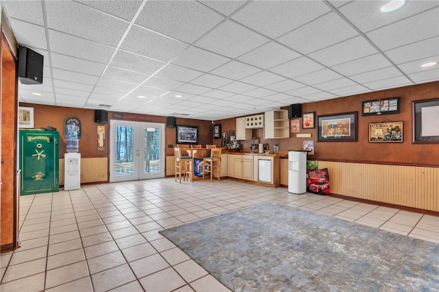 interior space with light tile patterned floors, french doors, a drop ceiling, and wainscoting