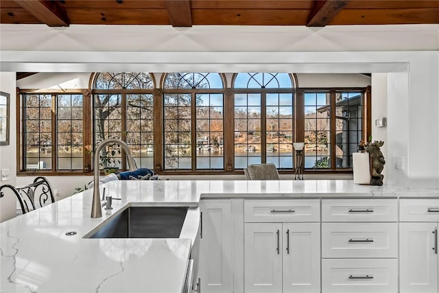 kitchen with light stone counters, beam ceiling, white cabinetry, and a sink