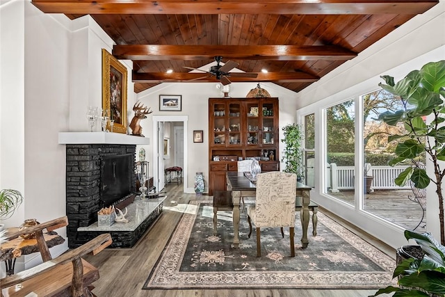 dining room featuring a ceiling fan, wooden ceiling, wood finished floors, vaulted ceiling with beams, and a stone fireplace