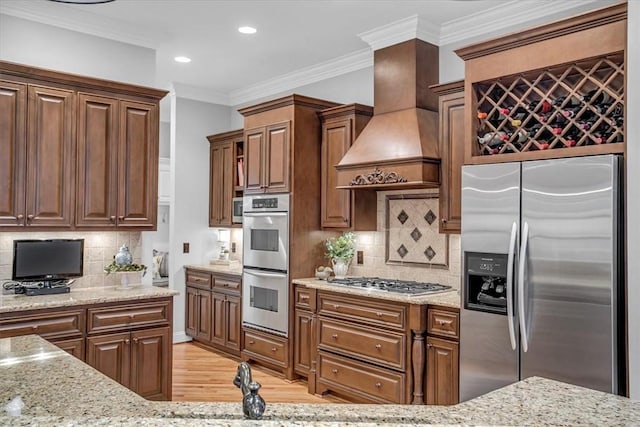 kitchen with stainless steel appliances, light wood-style floors, custom range hood, light stone countertops, and crown molding