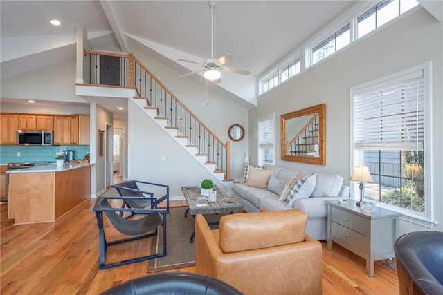 living area featuring ceiling fan, beamed ceiling, stairs, light wood-style floors, and high vaulted ceiling