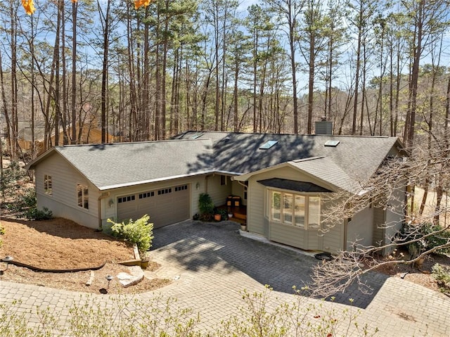 ranch-style house featuring an attached garage, a shingled roof, a chimney, and decorative driveway