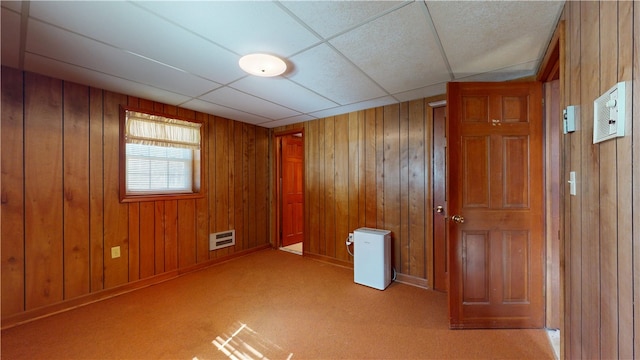 carpeted empty room featuring heating unit, wooden walls, and a drop ceiling