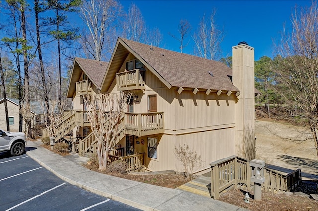 exterior space with a chimney, uncovered parking, a shingled roof, a balcony, and stairs