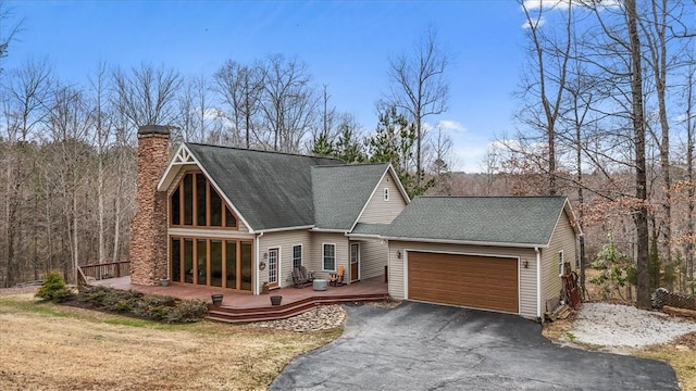 chalet / cabin featuring aphalt driveway, an attached garage, roof with shingles, a wooden deck, and a chimney