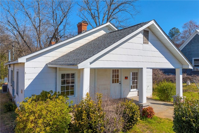 view of front facade featuring central air condition unit, covered porch, roof with shingles, and a chimney