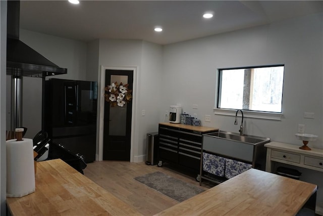 kitchen featuring wall chimney range hood, butcher block countertops, light wood-style flooring, freestanding refrigerator, and a sink