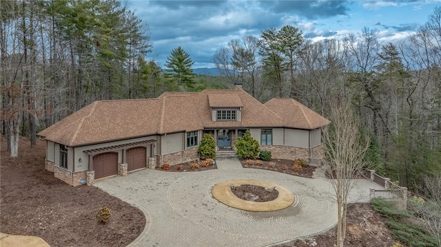 view of front of house with an attached garage, a chimney, curved driveway, stone siding, and a view of trees