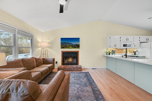 living area featuring baseboards, light wood finished floors, lofted ceiling, ceiling fan, and a brick fireplace