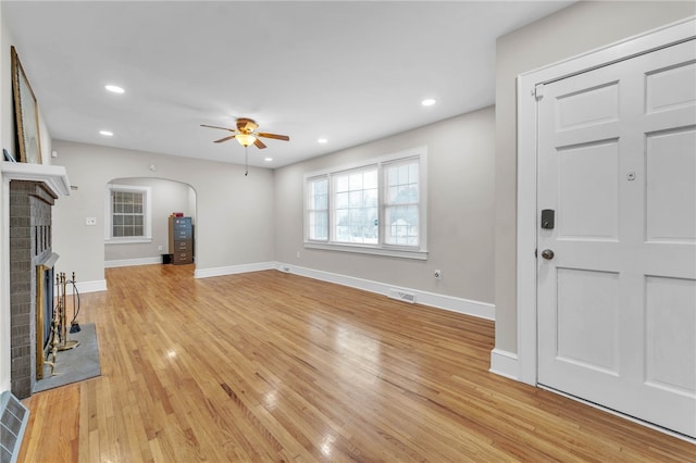 unfurnished living room with arched walkways, a brick fireplace, light wood-type flooring, and baseboards