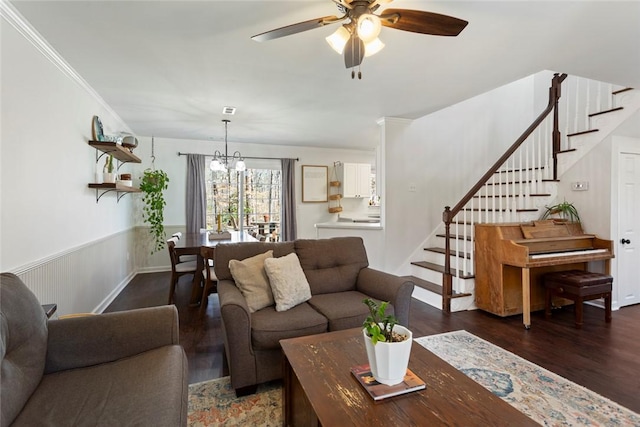 living area with a wainscoted wall, ceiling fan with notable chandelier, wood finished floors, crown molding, and stairs