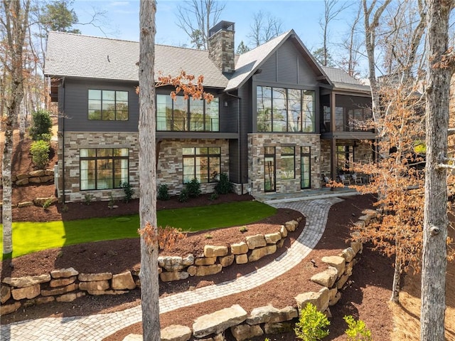 rear view of property featuring a lawn, stone siding, roof with shingles, and a chimney