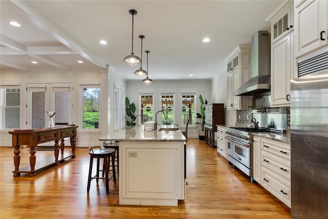 kitchen with light wood finished floors, a kitchen breakfast bar, high end appliances, wall chimney range hood, and an island with sink
