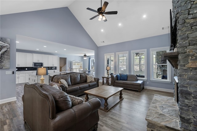 living room with dark wood finished floors, ceiling fan, high vaulted ceiling, and a stone fireplace