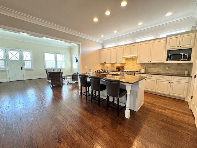 kitchen featuring a kitchen island with sink, dark wood-style flooring, under cabinet range hood, decorative backsplash, and ornamental molding