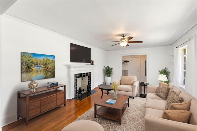living room featuring baseboards, ornamental molding, a tiled fireplace, a ceiling fan, and wood finished floors