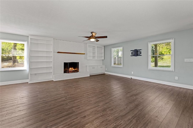 unfurnished living room with built in shelves, ceiling fan, baseboards, a brick fireplace, and dark wood-type flooring