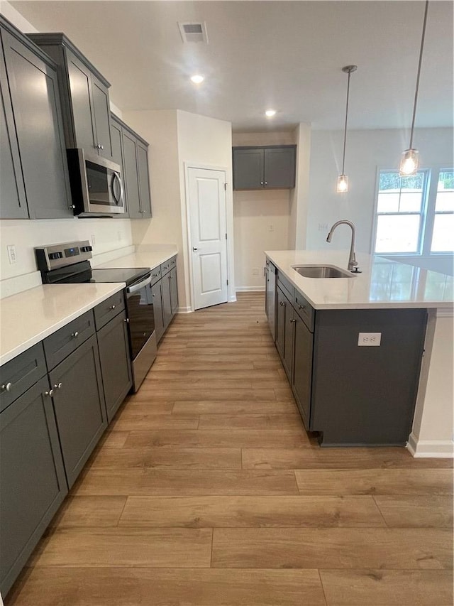kitchen with an island with sink, stainless steel appliances, a sink, light wood-style floors, and baseboards
