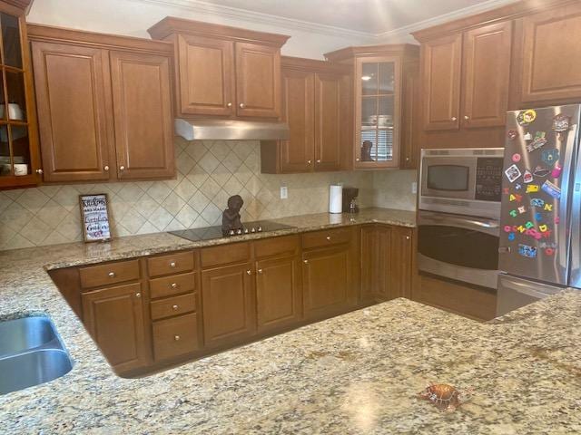 kitchen featuring stainless steel appliances, decorative backsplash, glass insert cabinets, light stone counters, and under cabinet range hood