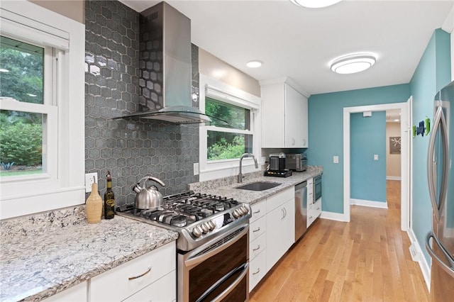 kitchen with wall chimney range hood, light stone counters, a sink, stainless steel appliances, and light wood-type flooring