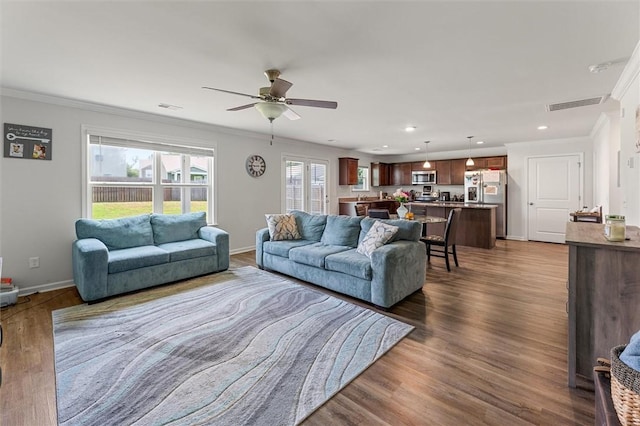 living area with a ceiling fan, visible vents, baseboards, ornamental molding, and dark wood finished floors