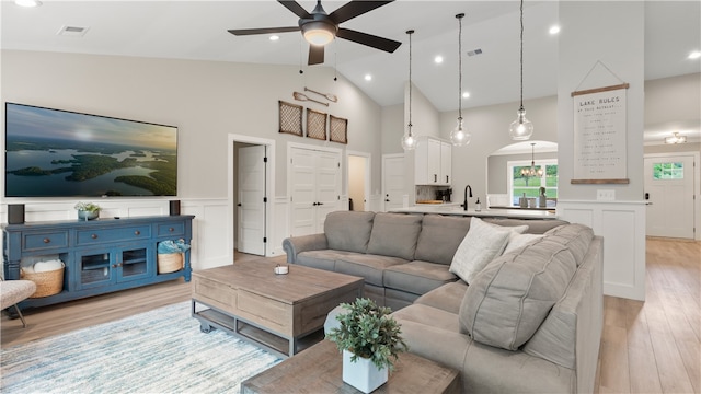living area featuring ceiling fan, a decorative wall, high vaulted ceiling, light wood-style flooring, and a wainscoted wall