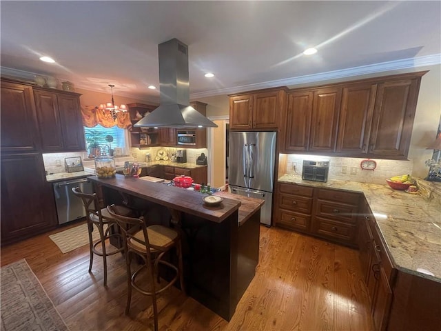 kitchen featuring island exhaust hood, appliances with stainless steel finishes, tasteful backsplash, light stone counters, and an inviting chandelier