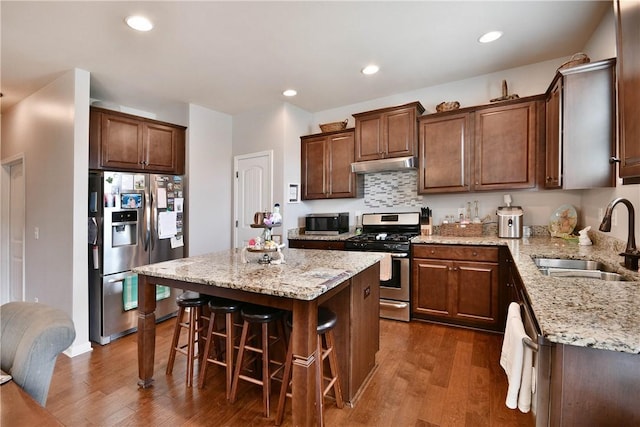 kitchen featuring appliances with stainless steel finishes, light stone countertops, dark wood finished floors, a sink, and under cabinet range hood