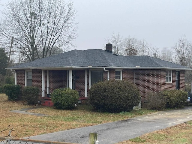 ranch-style house featuring covered porch, a chimney, roof with shingles, brick siding, and a front lawn