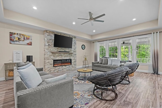 living area with wood finished floors, baseboards, a stone fireplace, ceiling fan, and a raised ceiling