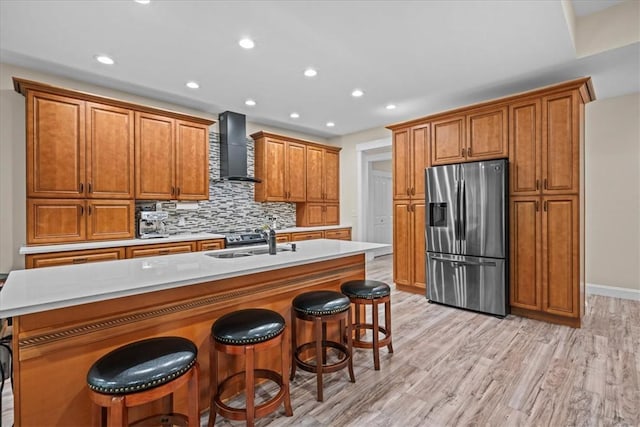kitchen featuring brown cabinetry, stainless steel refrigerator with ice dispenser, a sink, recessed lighting, and wall chimney exhaust hood